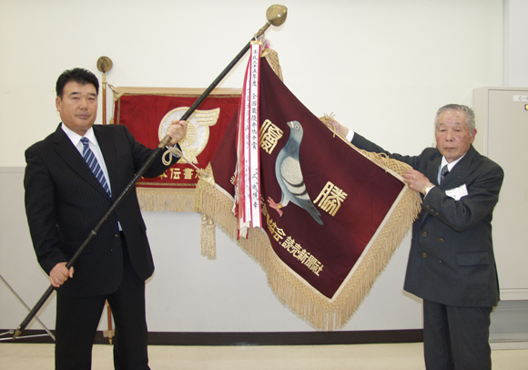 ▲全国最優秀鳩舎賞のシンボル「協会旗」を拡げる三宅博幸鳩舎(左)と藤井一一新会長。